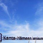 Kuvassa on Kanta-Hämeen keskussairalaan katolla oleva valomainos, jossa on sairaanhoitopiirin logo. Lisäksi kuvassa on nähtävissä sinistä taivasta ja pilviä